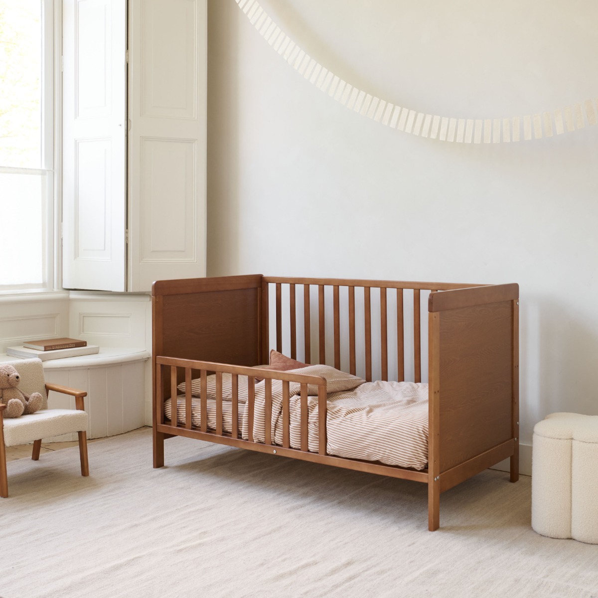 Lit bébé en bois couleur noyer avec barrière de sécurité, installé dans une chambre lumineuse au style doux et minimaliste.